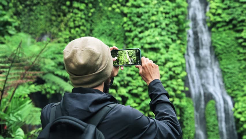 Traveling Man in Green Nature of Mountain Landscape with River Waterfall in Summer Forest. Unrecognizable Person Outdoors Taking Photo or Video of Water Fall View. Phone Photography for Social Media