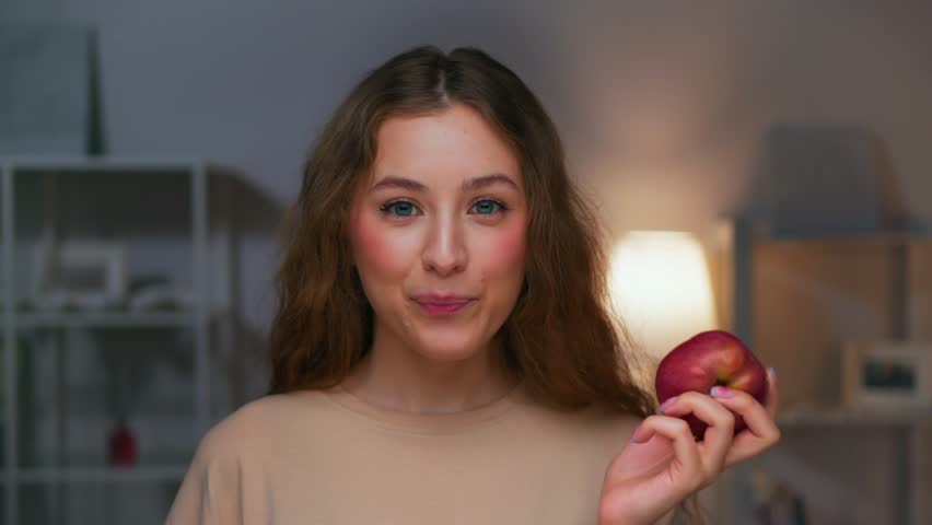 Portrait of Young Woman Eating Healthy Food and Looking at Camera. Real Person on Diet Consuming Fresh Red Apple as Lunch Snack. Playful Female Human Inside Cozy Living Room Alone with Organic Meal