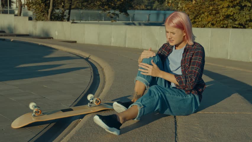 Upset trendy attractive pink haired female skateboarder holding and massaging injured knee, feeling pain and discomfort after fall while skateboarding on city street at sunrise. Royalty-Free Stock Footage #1109527395