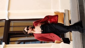 business lady teenager in black dress sunglasses and red jacket with handbag in hands. the girl stands on the sidewalk and crosses the street after a car passes by. vertical video - Powered by Shutterstock - Get 15% off with code: PIKWIZARD15