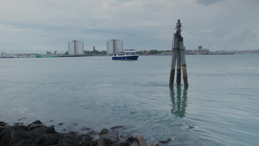Wide shot of seawater hitting rocks and post on a calm day in the harbour, a boat is passing by with council estates in the background. Handheld shot. 1080p.
