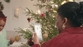 Young African American woman recording video on smartphone of her little daughter decorating Christmas tree with handmade toys in cozy domestic atmosphere