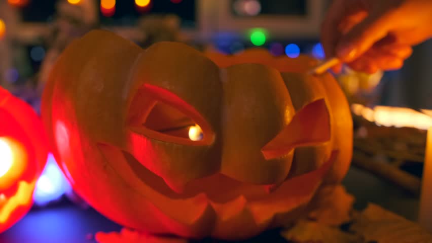 Traditional carved pumpkin, hand lights a candle jack o lantern lights up with flickering lights on blurred background glowing garlands at night festive background Halloween holiday, 31 October. Royalty-Free Stock Footage #1109537945