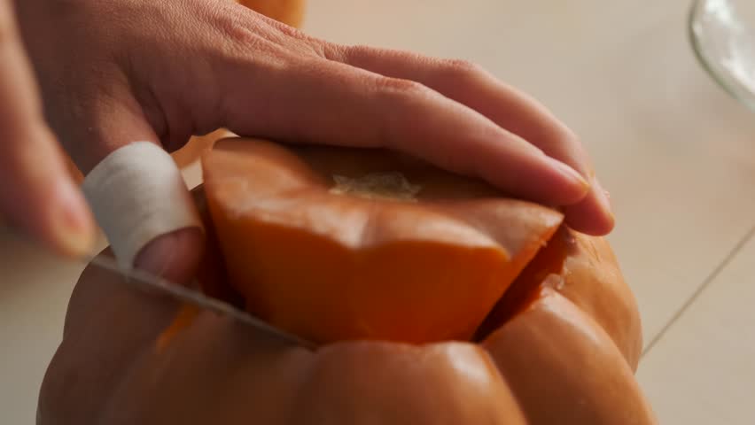 Сlose-up of pumpkin and woman's hands with bandaged finger opening  carved lid to remove flesh and peel sweet vegetable preparing it for cooking and carving jack-o'-lantern. During Halloween.