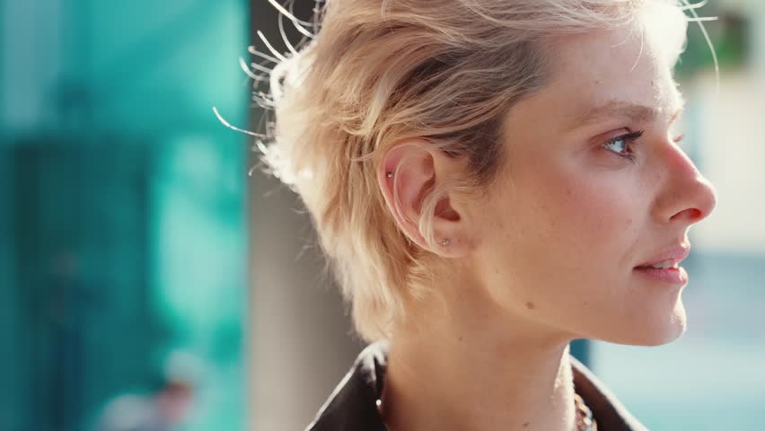 Portrait of short-haired blonde female with confident look and natural beauty turning eyes at camera and making gentle smile. Gorgeous woman with necklace on neck having walk on city streets. Royalty-Free Stock Footage #1109539591