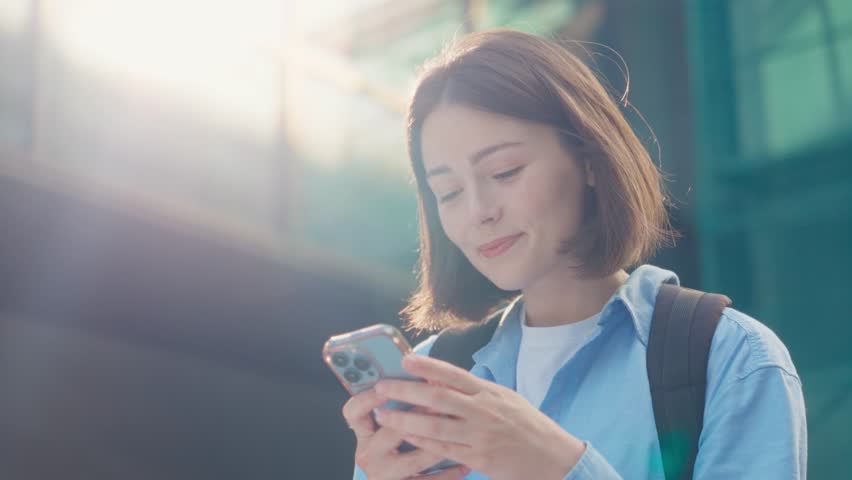 Short-haired brunette in casual wear standing near large commercial building and looking at cellphone screen. Smiling lady browsing internet and reading exciting news online. | Shutterstock HD Video #1109539597