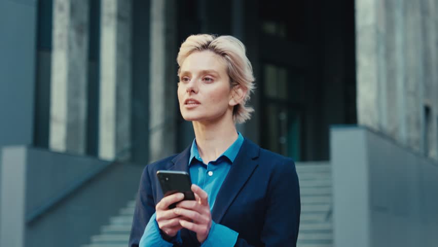 Elegant female blonde dressed in formal attire using modern cell phone while walking outdoors. Caucasian business lady chatting with coworkers online outside office center. | Shutterstock HD Video #1109539599