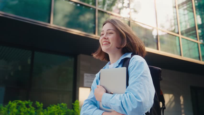 Cheerful young lady with short hair carrying portable laptop in hands and happily walking on urban street. Casually dressed positive student with backpack attending classes. | Shutterstock HD Video #1109539607