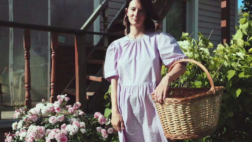 portrait of a brunette woman in a lilac dress in the garden near the house with a basket of harvest against the background of bushes of hydrangea flowers