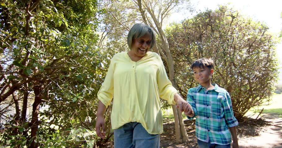 Happy biracial grandmother and grandson walking in sunny garden, slow motion. Family, togetherness, nature and lifestyle, unaltered.