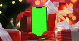 Christmas gift phone with green screen, close up. Smartphone with mockup touchscreen in red presents on glowing New Year garland backdrop. Static empty advertising space on bokeh gold lights backdrop