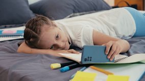 Pre-teen kid watching funny videos on smartphone while resting on bed during break from doing homework in room after school