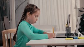 Pre-teen female kid greeting her teacher during online lesson on laptop while sitting at desk and writing in copybook at home