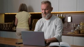 Senior Caucasian family in kitchen mid aged man husband retired businessman remote working distance learning online from home using laptop computer while mature woman wife wash dishes household work - Powered by Shutterstock - Get 15% off with code: PIKWIZARD15