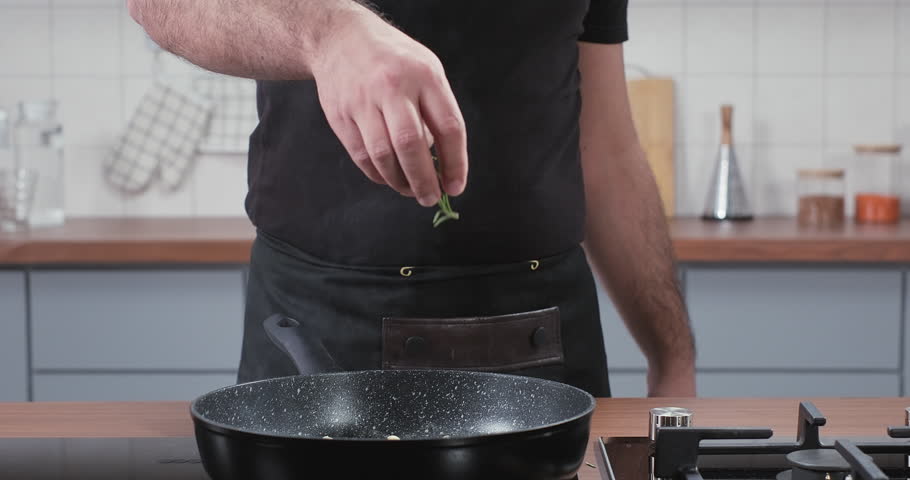 Chef throws rosemary into the steaming frying pan. Adding fresh rosemary herbs in slow motion. Cooking at home