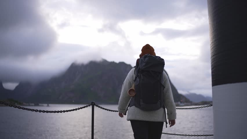 The enchanting scene unfolds in slow motion as a young woman gracefully makes her way to a white lighthouse in Reine, Lofoten, Norway.