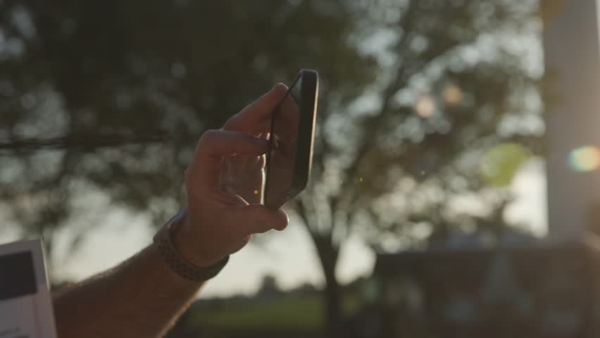 Slow motion of middle-aged man with hairy arms taking photo on smart phone in a public park near downtown Washington D.C. with sun flares, trees, pedestrians, and greenery during autumn