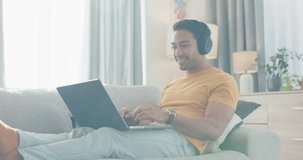 Man, computer and headphones on sofa listening to music, e learning and relax in online education or subscription at home. Happy young asian student for audio streaming or search kpop video on laptop