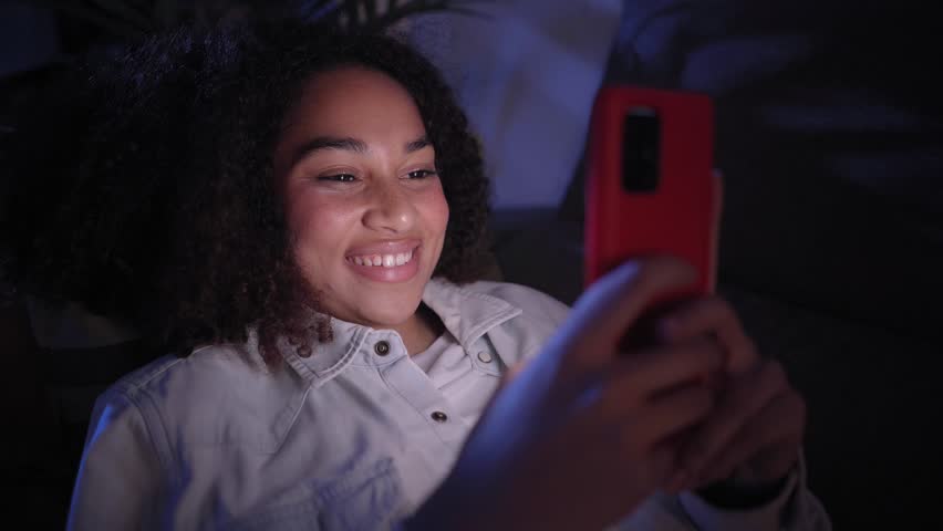 Close up of cheerful young latina girl using smart phone lying on sofa. Smiling woman chatting on mobile at home at night. Single people, dating apps and social networks in generation z. Royalty-Free Stock Footage #1109561767