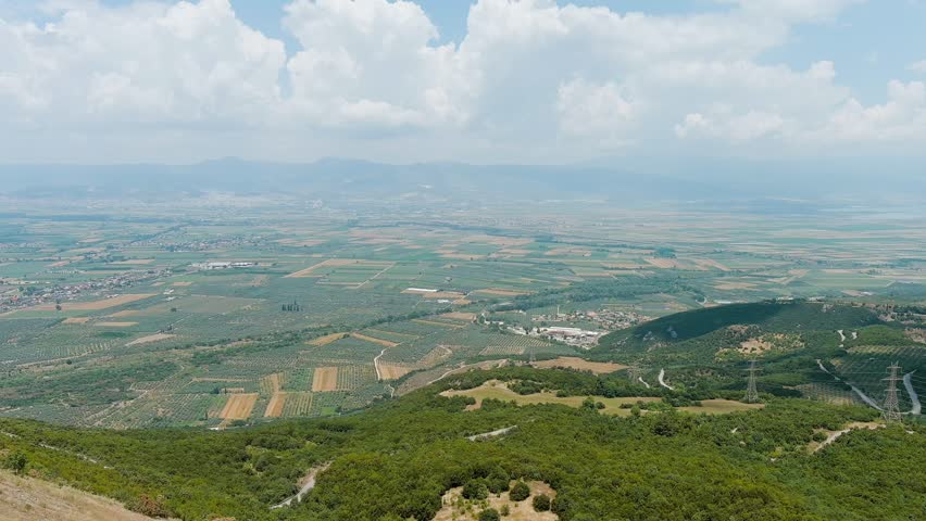 Lamia, Phthiotis, Greece. A sweeping aerial panorama of the Spercheios Valley reveals an expansive patchwork of agricultural fields and olive groves stretching toward the horizon, framed by forested hills in the foreground and hazy mountain ranges in the distance. The landscape is divided into geometric parcels of varying greens, golds, and earth tones, interspersed with winding dirt roads and small clusters of rural buildings. The valley, described by Homer as having rich alluvial soil, has been a center of cultivation since antiquity. Summer clouds drift across a bright sky, casting soft shadows over the terrain. The camera maintains a steady elevated perspective, capturing the scale and tranquility of this fertile Mediterranean plain. The scene conveys rural serenity, agricultural heritage, and the timeless relationship between the land and its cultivators.