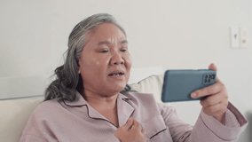Medium close up of sick Asian woman videocalling somebody on smartphone while resting in bed at home