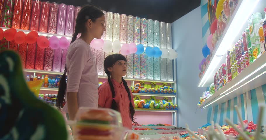 Two girls with brown hair braided and wearing a pink shirt choosing and buying candies in a store. Royalty-Free Stock Footage #1109571715