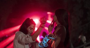 Two happy young girls dancing on open space party with flashing lights and lasers. Out of focus crowd and dj stage in the background.