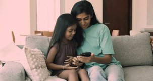 Beautiful Indian woman spend time with cute little 5s daughter sit on sofa with modern smart phone, enjoy new amusing mobile application, teach kid how to use gadget, make call, having fun on internet