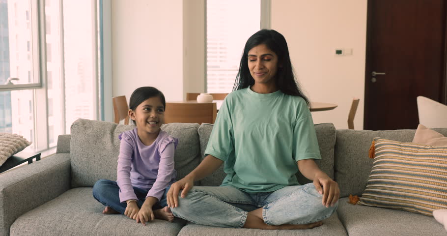 Little playful girl and her young mom meditating together seated on sofa in living room at home. Loving mother teach yoga practice daughter. Lifestyle, good life habit of Indian family, mindfulness Royalty-Free Stock Footage #1109599921