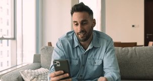 Hispanic man sit on couch at home holding smartphone make video call to friend or family living abroad, look at device screen, make speech, vlogger share information with followers using videocall app