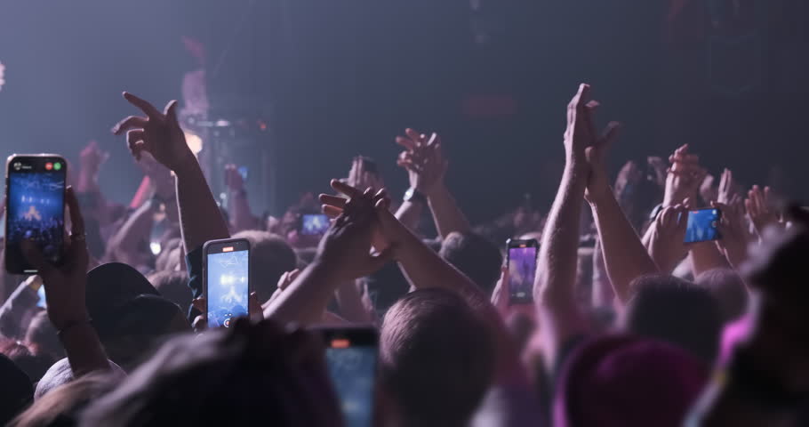 Waving fans hands at band concert in magenta stage light spotlights. Close up, happy people clap their hands, film show on phones. Disco party illumination by pink orange light. Crowd audience dance