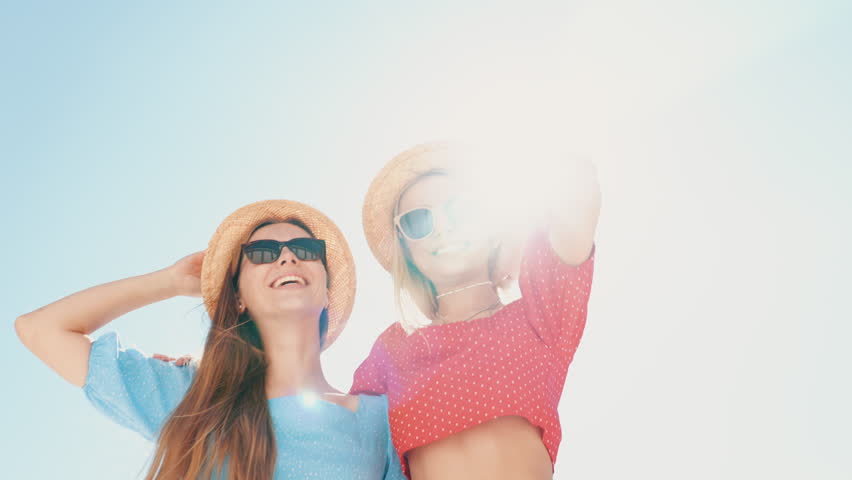Two young beautiful smiling hipster women in trendy summer same clothes. Carefree models posing in the street. Positive models having fun. Cheerful and happy, at sunny day. In hat and sunglasses Royalty-Free Stock Footage #1109605413