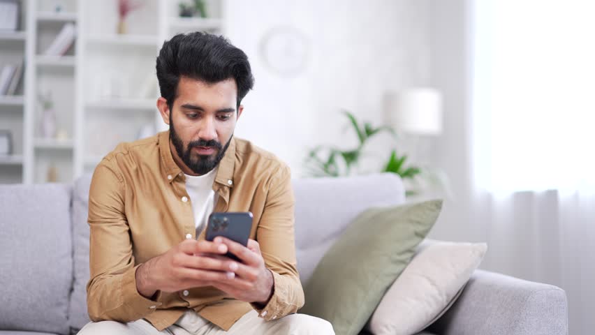 Young bearded man using smartphone sitting on sofa in living room at home. Smiling handsome male browses social networks, does shopping in online store, chats with a friend, writes or reads messages Royalty-Free Stock Footage #1109605737