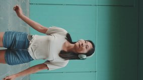 Vertical video: Happy beautiful brunette girl in headphones dressed in casual clothes, listens to music and dances on blue wall background