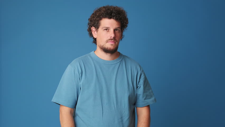 Happy guy with curly hair dressed in blue t-shirt looking at camera and showing sign of shape heart isolated on blue background in studio