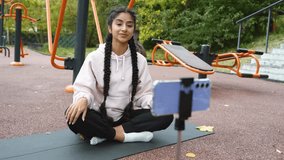 Online fitness training in modern outdoors sportground performed by young Indian woman looking at smartphone