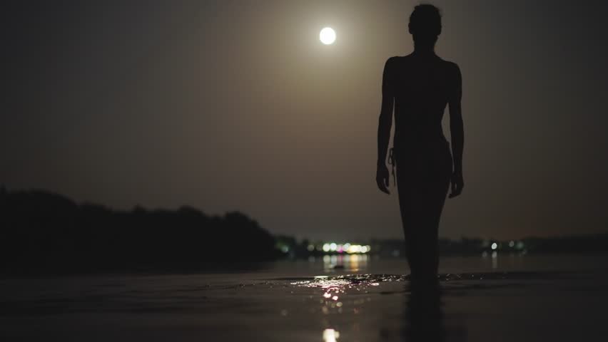 silhouette of slim woman in bikini walking in shallow water in lake at night with moonlight. summertime night sensual girl swimming in darkness