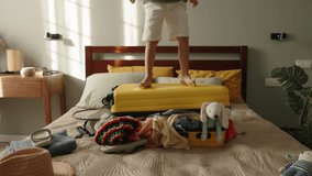 Boy joyfully jumps on packed suitcase. Small child traveler tourist, enjoys summer holidays, jumps, rejoices, dances on baggage on bed among messy clothes. Overflowing with joy fun happiness delight  - Powered by Shutterstock - Get 15% off with code: PIKWIZARD15