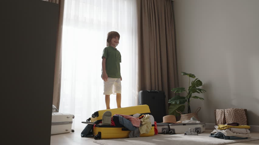 Boy joyfully jumps on packed suitcase. Child happily jumps on chaotically messy self-packed suitcase filling air with fun. Diligent little child toddler boy rejoices after packing clothes in suitcase Royalty-Free Stock Footage #1109627939