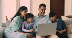 Happy Indian family with little adorable son and daughter relaxing at home, watching online movie, spend free time together, enjoy cartoons seated on cozy couch with laptop. Family leisure on internet