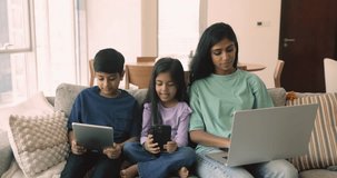 Young woman their little kids using diverse electronic devices seated on sofa at home. Family spend free time on internet, use tablet, laptop and cellphone for fun and communication. Gadget addiction