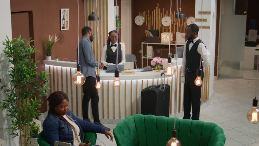 Tourist calls for assistance and room card at reception using front desk bell to talk to employee in lobby. African american hotel guest with suitcases receiving access key for accommodation.