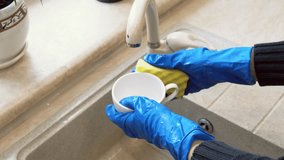 Cropped video of young woman is washing coffee cup using a sponge while doing cleaning at home. Close-up of Housewife hands in blue rubber gloves washing dishes in the kitchen sink. 4K footage 25FPS