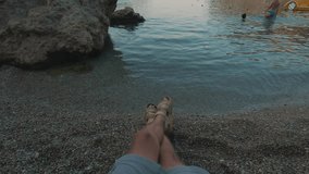 First person view - young man relaxing on pebble beach, looking at sea and putting on sunglasses, POV shot, Mediterranean sea, Greece