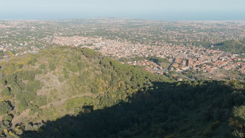 Nicolosi, Sicily, Italy. Volcanic craters densely overgrown with forest on the southern slopes of Mount Etna, captured in a steady aerial view. The scene shows twin pyroclastic cones blanketed in green woodland, rising above the surrounding terrain with the town of Nicolosi and the distant coastline visible in the background. These are the Monti Rossi, formed during the devastating 1669 eruption that sent lava flowing toward Catania. Today the craters are cloaked in pine forest and Mediterranean vegetation, creating a striking contrast between dormant volcanic geology and thriving woodland. The camera maintains a broad elevated perspective, revealing the texture of the forested cones, the urban spread below, and the hazy blue line of the sea on the horizon. Bright daylight and clear atmospheric conditions emphasize the natural colors and topographical detail of this iconic Sicilian landscape.
