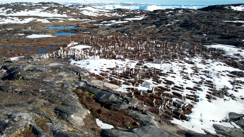 Dramatic aerial view over white crosses, grave stones in small cemetery to bay of melting icebergs. Greenland