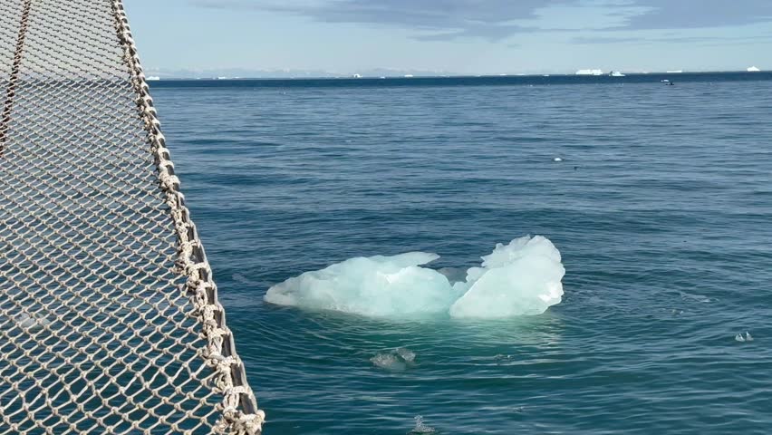 A piece of ice floats calmly in the sea water near a sailing ship. Scoresbysund, Greenland.