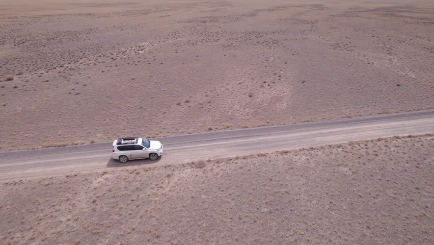 A white SUV is driving fast on a dusty road in the steppe. Clouds of dust fly from under the wheels and from the roof of the car. The jeep easily passes a dirt road. Blue sky and white clouds.
