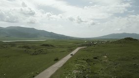 Drone video. Car driving on an old mountain road between green stony hills covered with grass. Ahead of the village, high mountains with rocky peaks in sunny weather. Armenia.