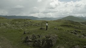 Drone video. Flyover around standing Caucasian man in jeans white t-shirt and black cap. Green stony hills covered with grass in sunny weather. Armenia.
